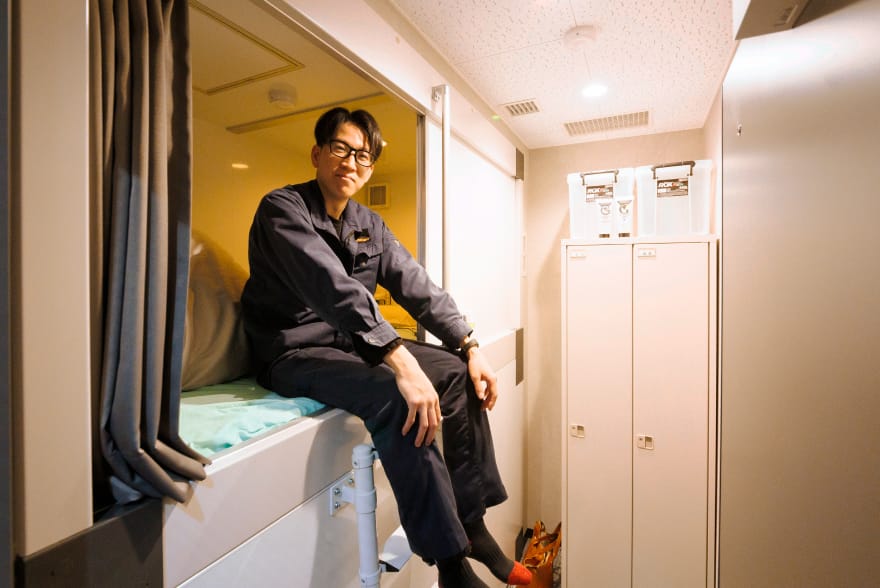 作業服を着た男性が簡易ベッドの上に座っており、仮眠室のような休憩スペースでくつろいでいる