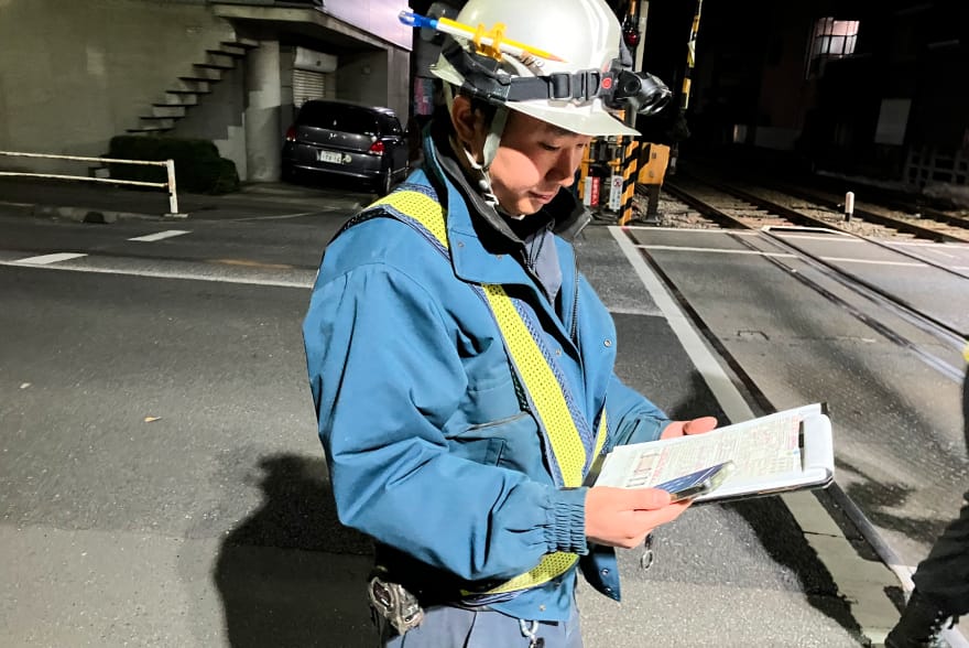 踏切付近でヘルメット姿の作業員が手帳とスマートフォンを使って点検記録を確認している