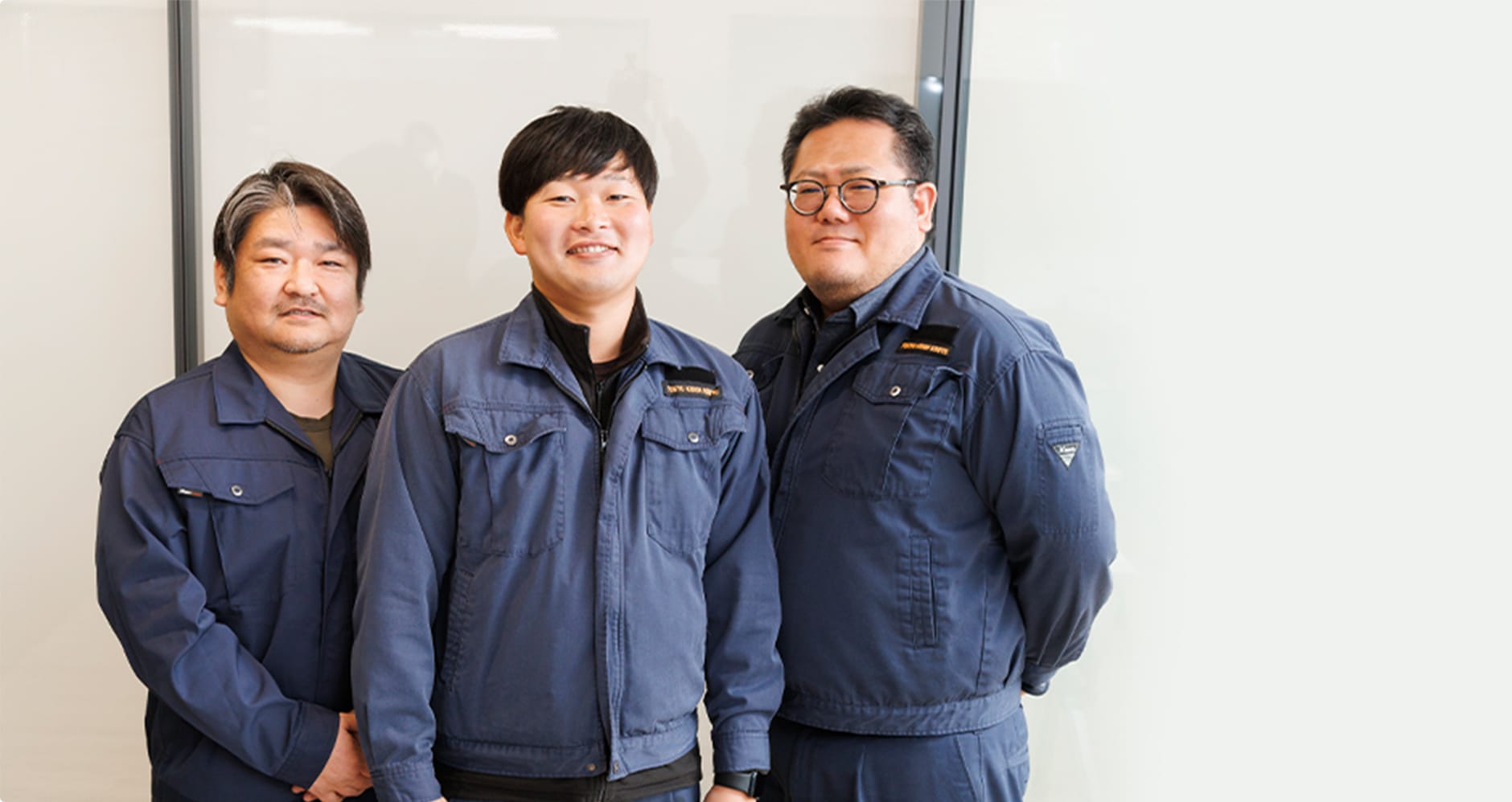 作業服を着た3人の男性が並んで立ち、カメラに向かって笑顔を見せている